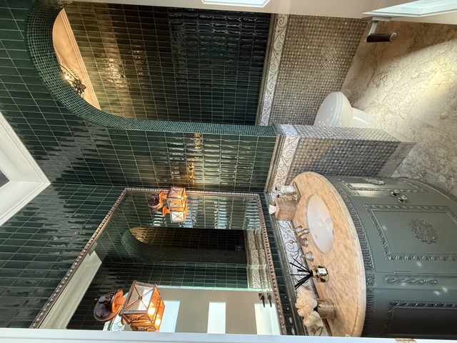 Elegant powder bath with glossy deep green tile, arched shower opening, custom painted vanity, stone countertop, mirror, and lantern sconces